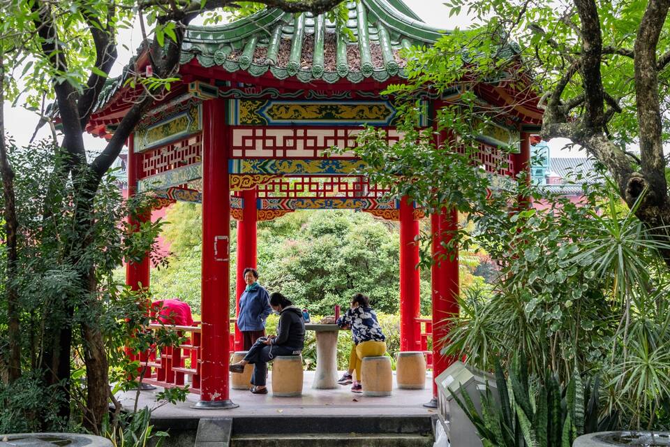 A traditional red and green pavilion with intricate designs, surrounded by lush greenery and trees.