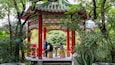 A traditional red and green pavilion with intricate designs, surrounded by lush greenery and trees.