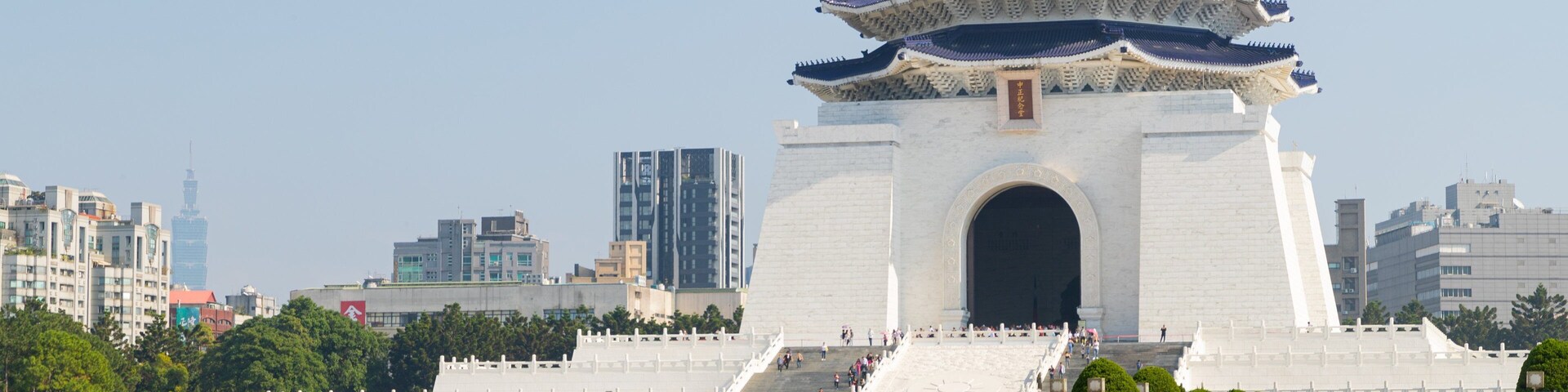 Um pavilhão em estilo tradicional chinês, com telhado azul e paredes brancas, rodeado por jardins bem cuidados e uma paisagem urbana ao fundo.