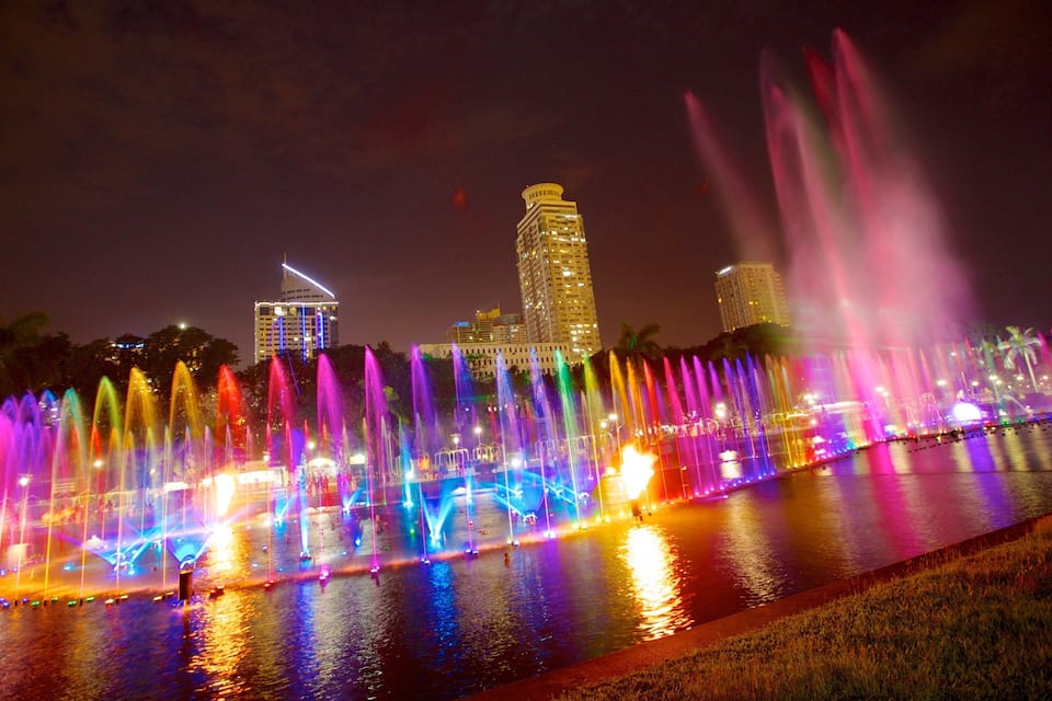A night-time water fountain show with colorful lights and a city skyline in the background.