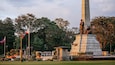 A monument with a tall obelisk and statues, surrounded by trees and potted plants.