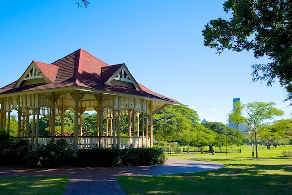 A gazebo with a red roof and white columns in a park with green trees and a clear sky.