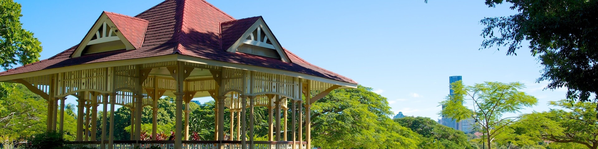Um gazebo com telhado vermelho e colunas brancas em um parque com árvores verdes e céu claro.