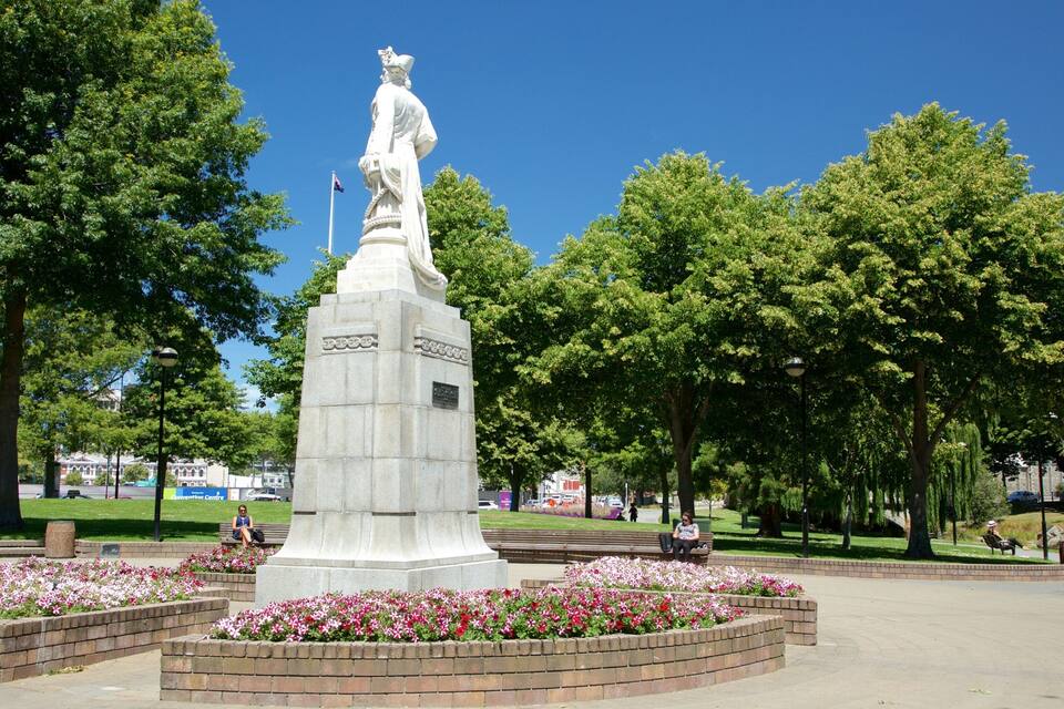 Uma estátua em um parque, cercada por canteiros de flores e árvores.