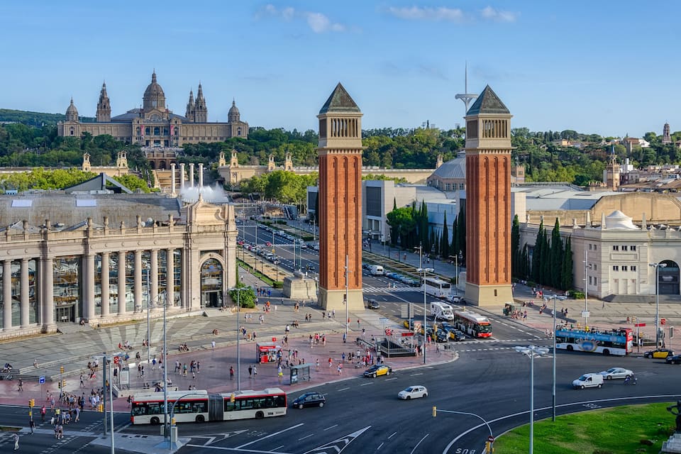 Un paysage urbain caractérisé par d’imposantes tours, un grand bâtiment orné de colonnes et une intersection animée où circulent des autobus et des voitures.