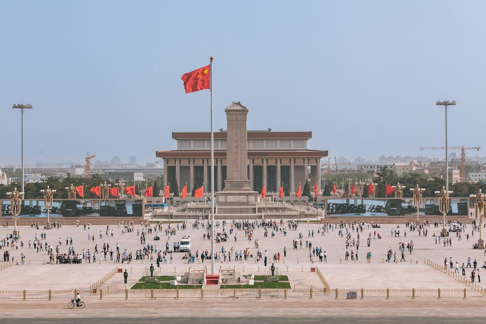 Une vaste place ouverte, ornée d’un monument imposant, de nombreux drapeaux rouges et d’un mât sur lequel flotte un drapeau rouge.