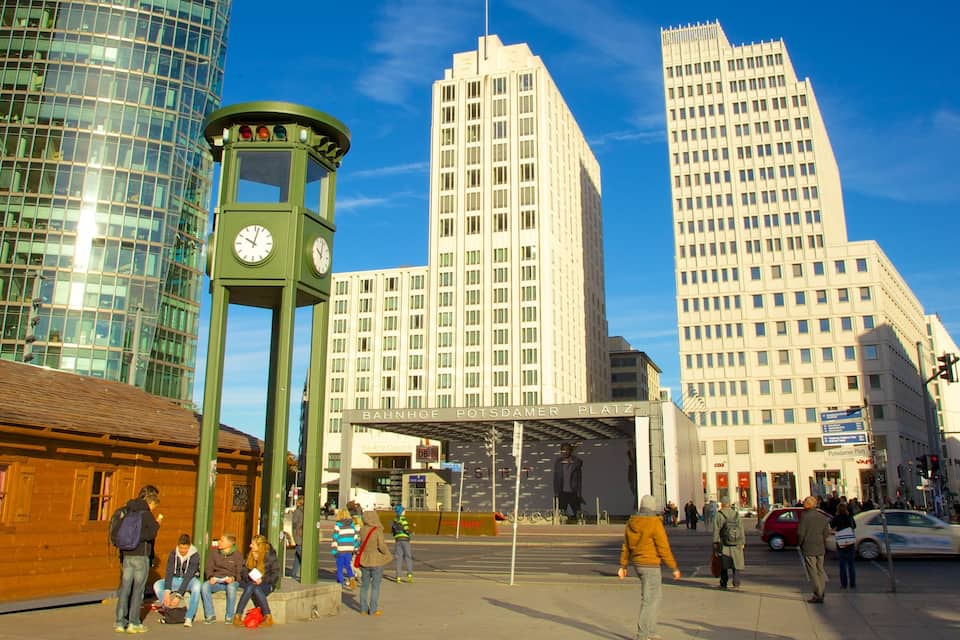 Una plaza urbana con una torre del reloj verde, edificios modernos y gente caminando.