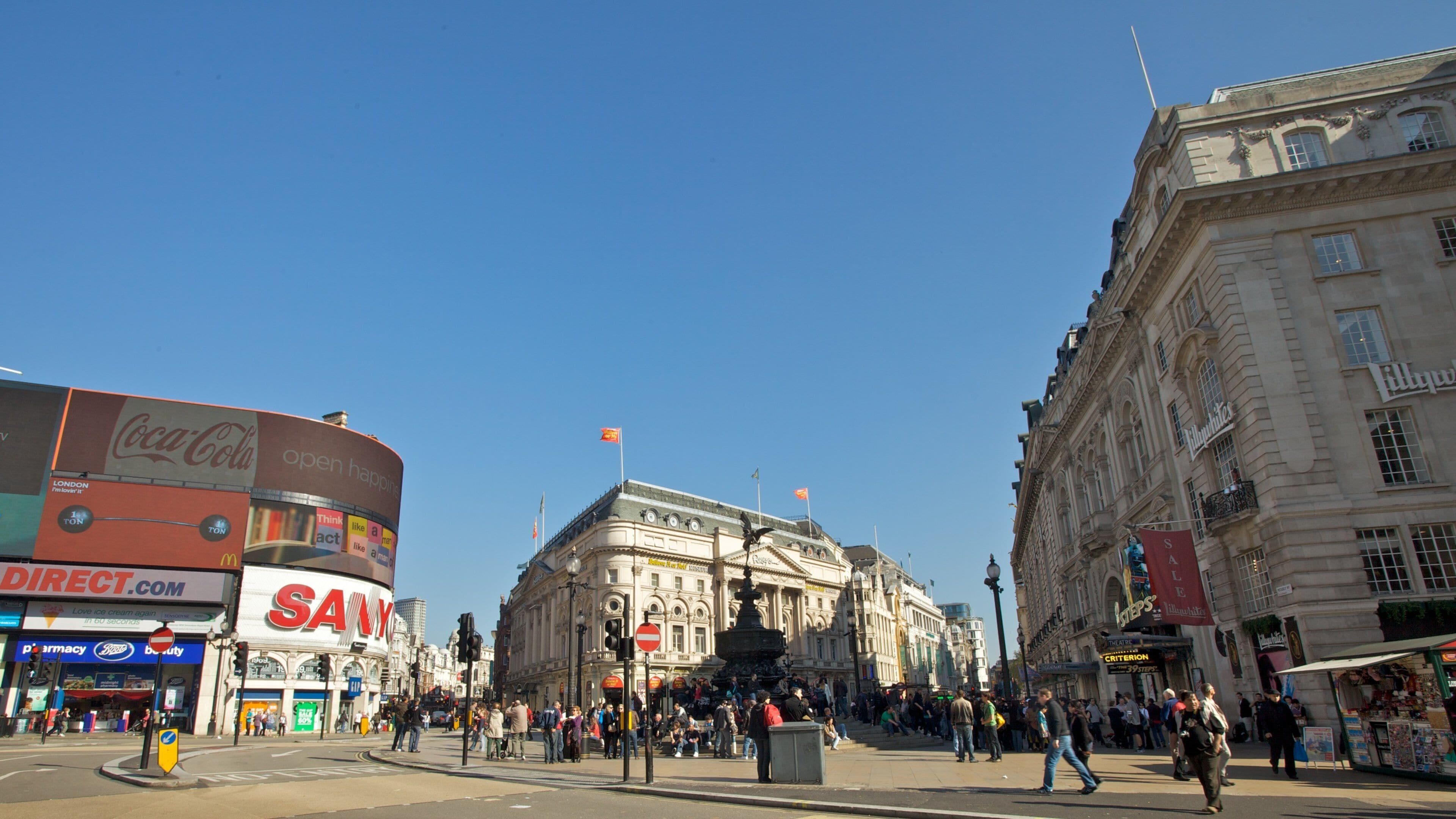 A busy city square with historic buildings, a fountain, and various shops including a pharmacy and a cinema.