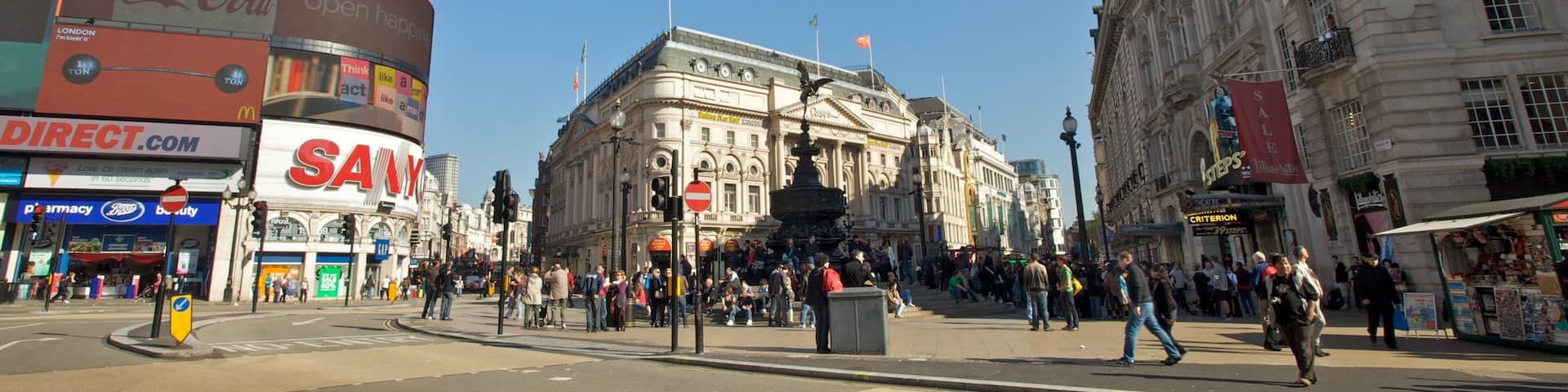 A busy city square with historic buildings, a fountain, and various shops including a pharmacy and a cinema.