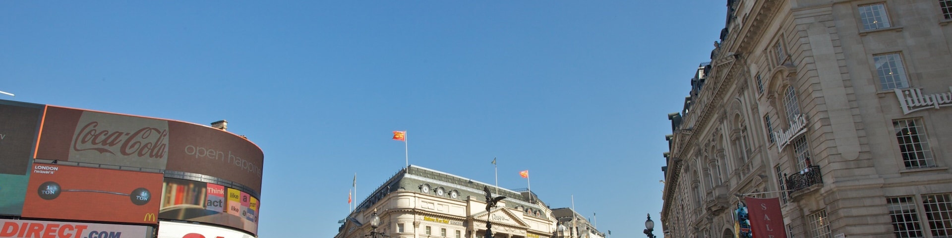 Uma praça movimentada na cidade, com edifícios históricos, uma fonte e diversas lojas, incluindo uma farmácia e um cinema.