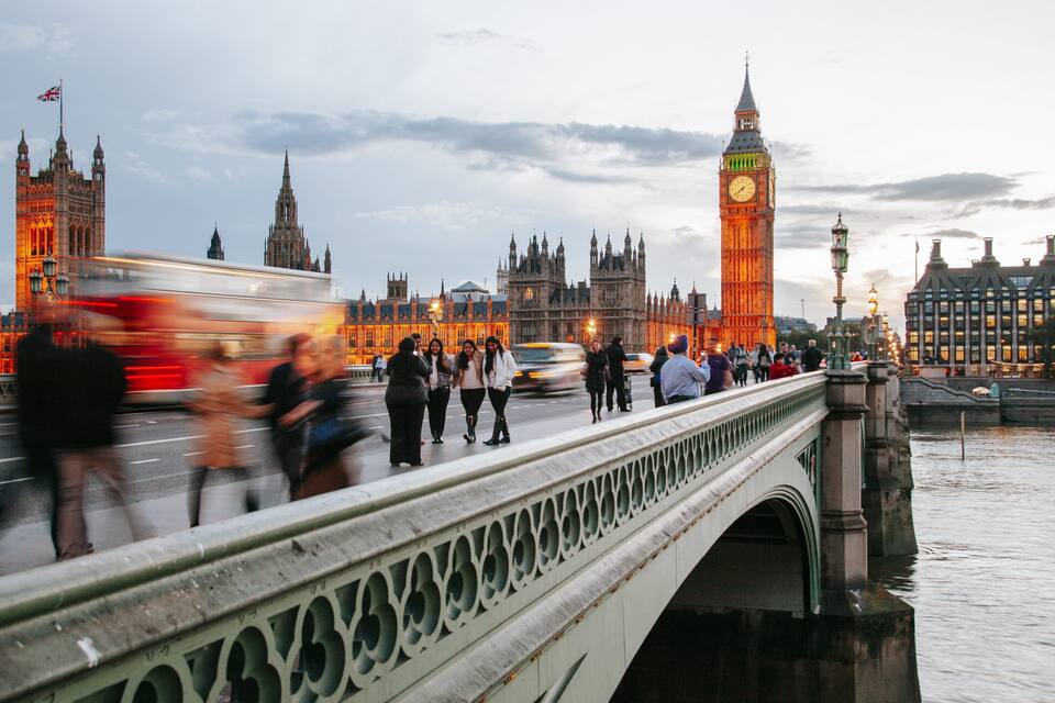 A bridge with people walking, a red double-decker bus, and the Big Ben clock tower in the background.