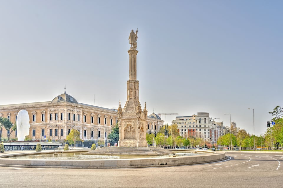 A large statue in a plaza with a fountain, surrounded by buildings.