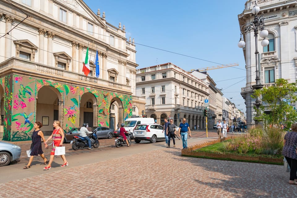 A cobblestone street lined with historic buildings, a mural with colorful floral designs, and flags of Italy and the European Union.