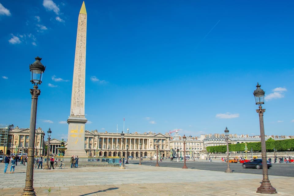Um obelisco alto numa praça com edifícios históricos e postes de iluminação.