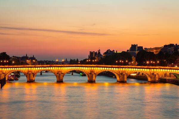 A lit stone bridge over a river at dusk with city buildings in the background.