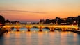 Un pont de pierre illuminé enjambe une rivière au crépuscule, avec des bâtiments de la ville en arrière-plan.