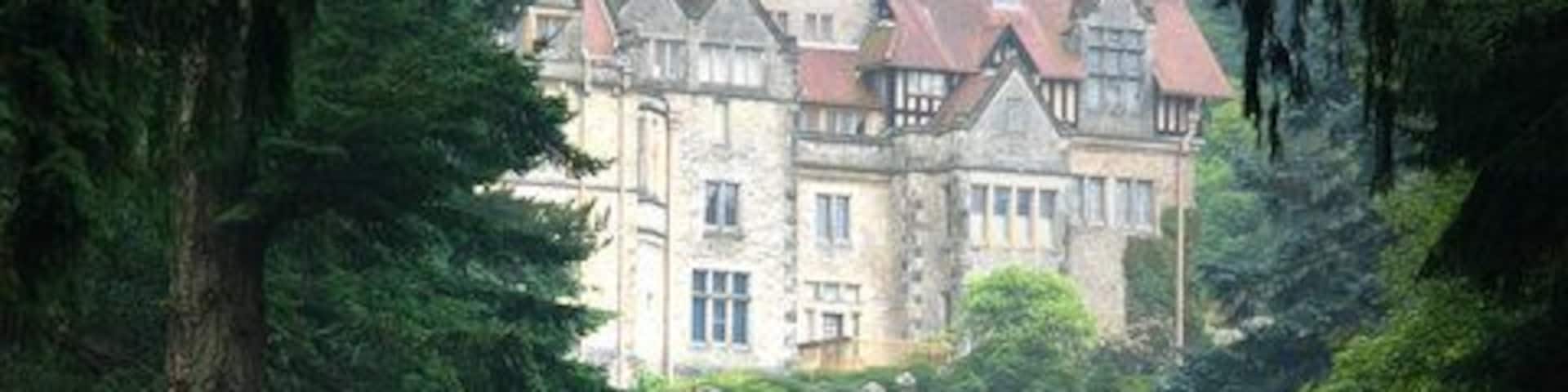 A large, historic house surrounded by dense forest and a stone bridge.