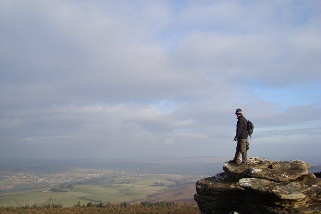 A person standing on a rocky outcrop with a panoramic view of a valley and distant cityscape.