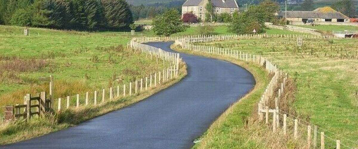 A narrow country road winding through a rural landscape with a fence on one side and a building in the distance.