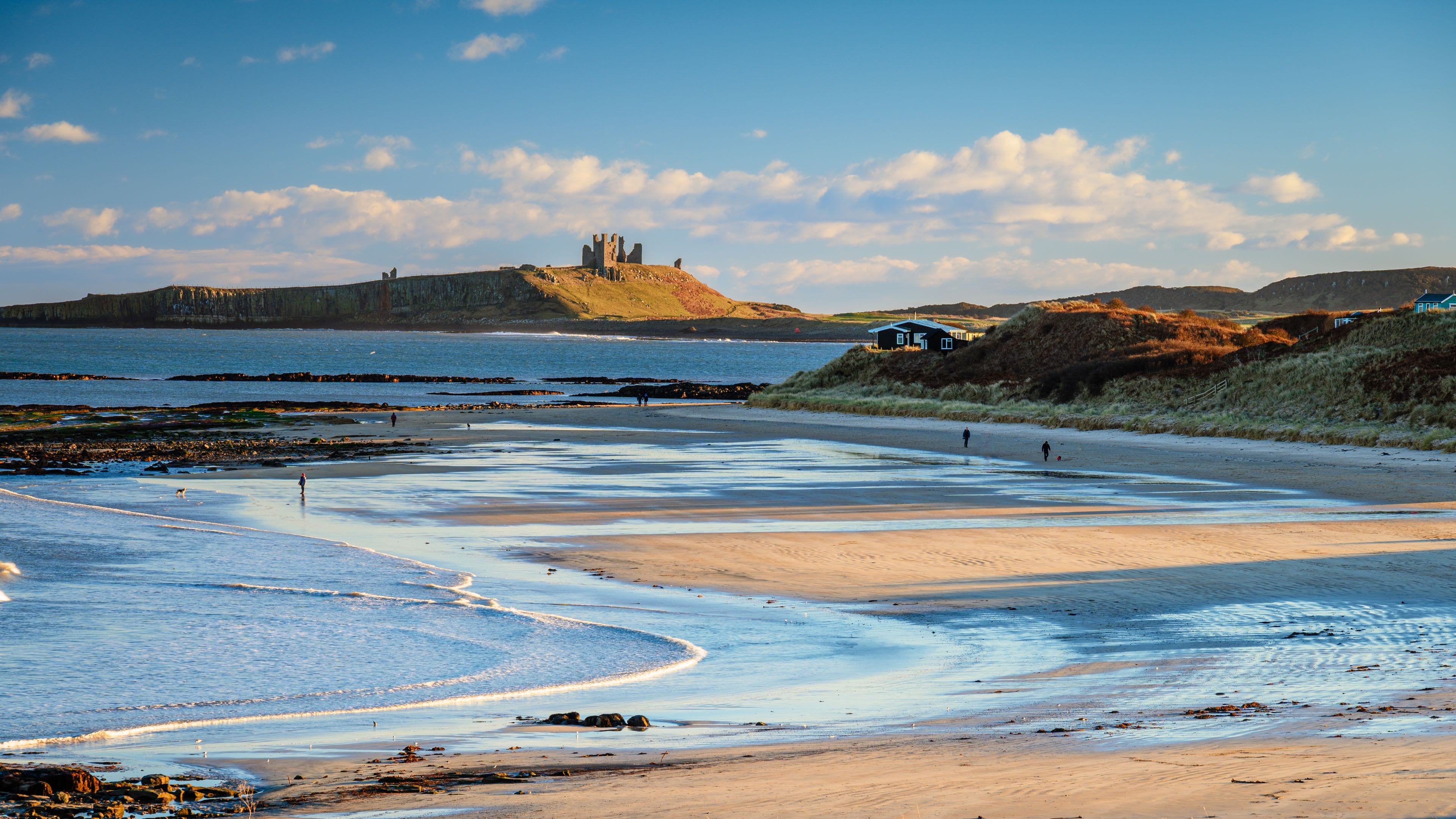 A beach with a castle ruin, calm sea, and grassy dunes.