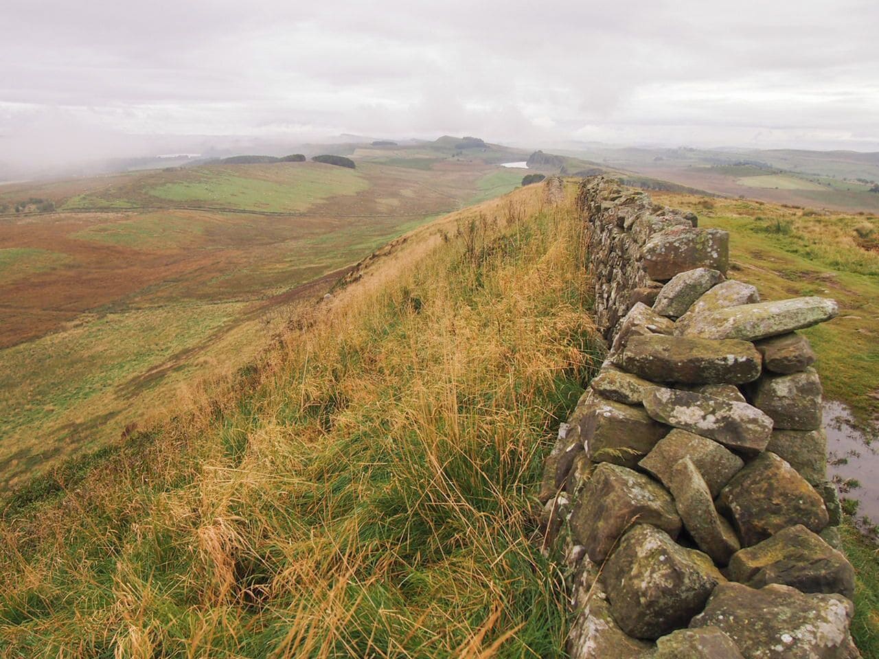 A stone wall on a hillside overlooking a valley with rolling hills and a cloudy sky.