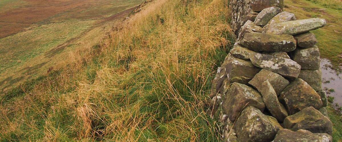 A stone wall on a hillside overlooking a valley with rolling hills and a cloudy sky.
