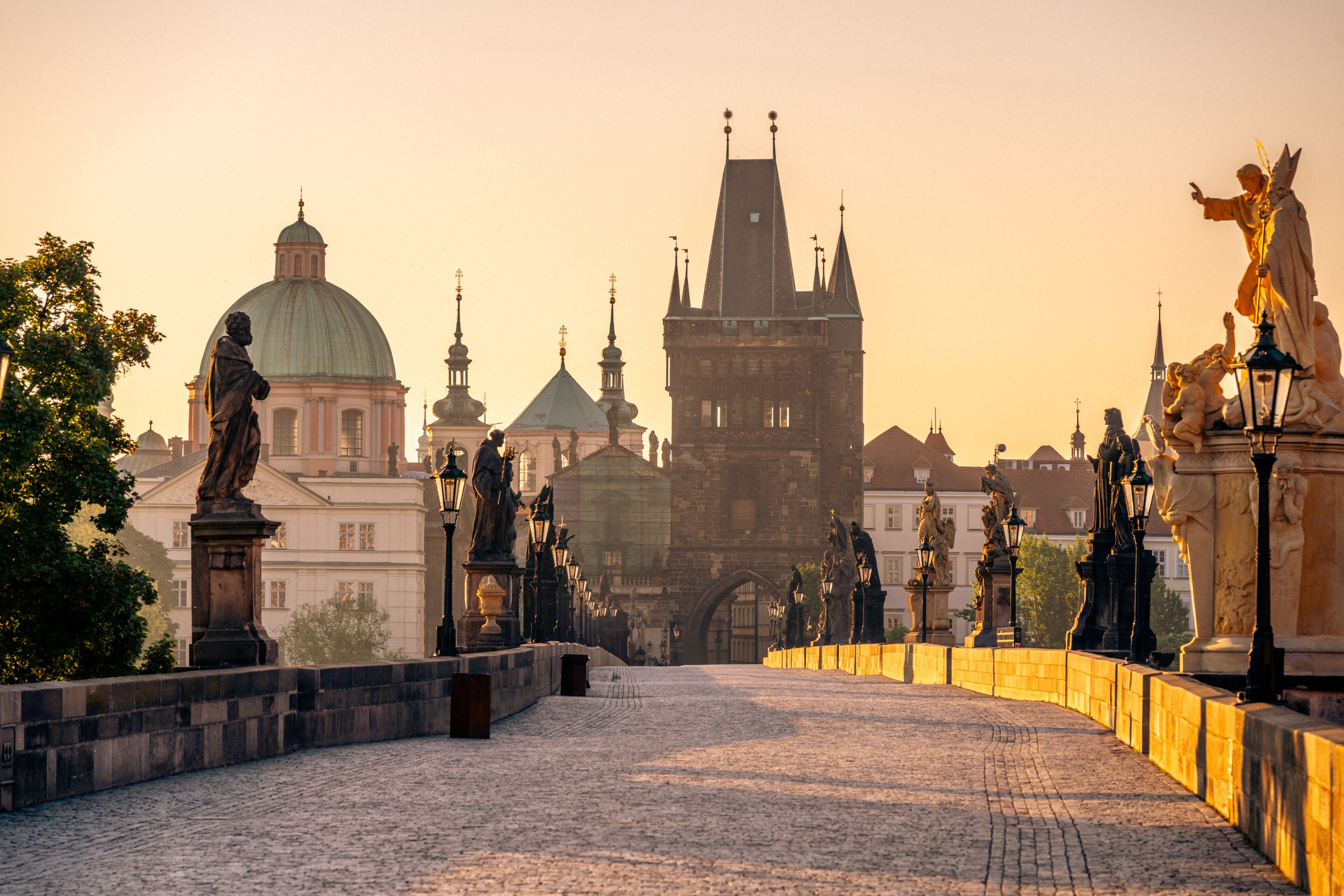 Sunrise casts a warm glow over Charles Bridge in Prague, highlighting the statues and the historic architecture. The Vltava River glistens, creating a serene morning atmosphere.