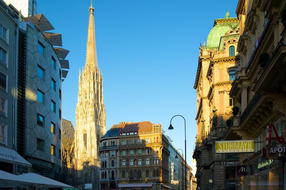 A city street with historic buildings, a cathedral spire, and storefronts including a pharmacy.
