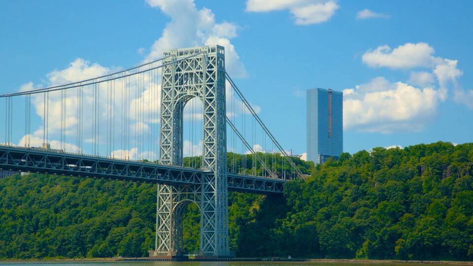 Uma grande ponte suspensa com estrutura de treliça de aço, um morro coberto por densa vegetação e um arranha-céu moderno ao fundo.