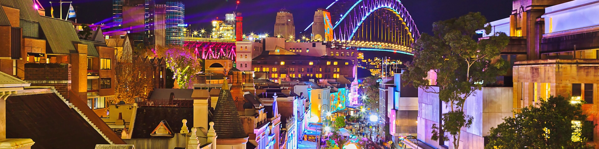 Un paisaje urbano nocturno con edificios iluminados, un puente destacado y una calle con tiendas.
