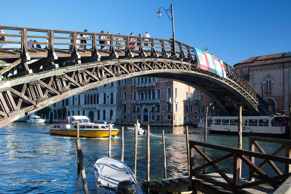 A wooden bridge over a canal with boats and a historic building in the background.