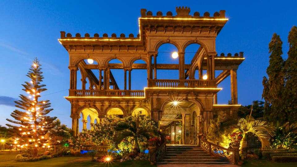 A grand, illuminated building with arched windows and a balcony, surrounded by lush greenery and decorative lights.