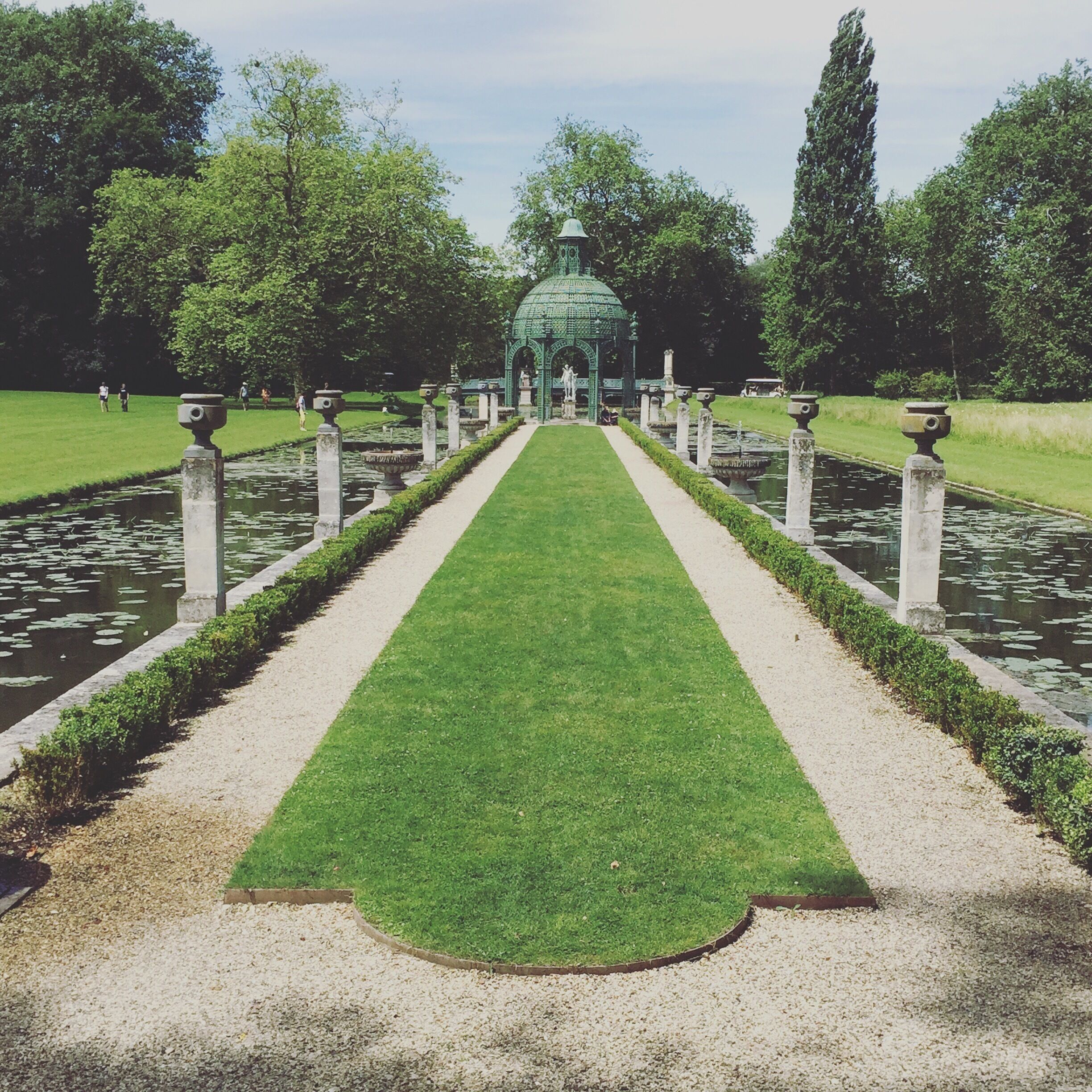 A formal garden with a central pathway, water features, and a green glass structure.