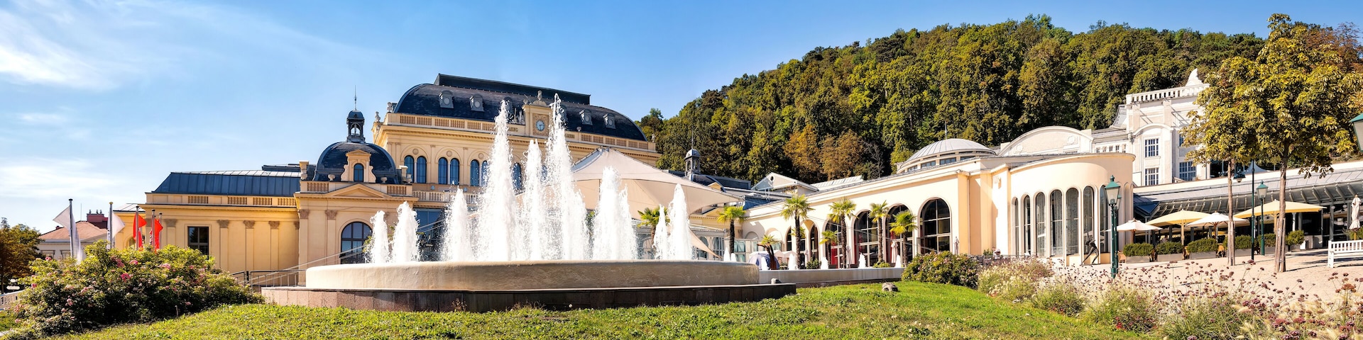 A historic building with a fountain, surrounded by lush greenery and a forested hill.