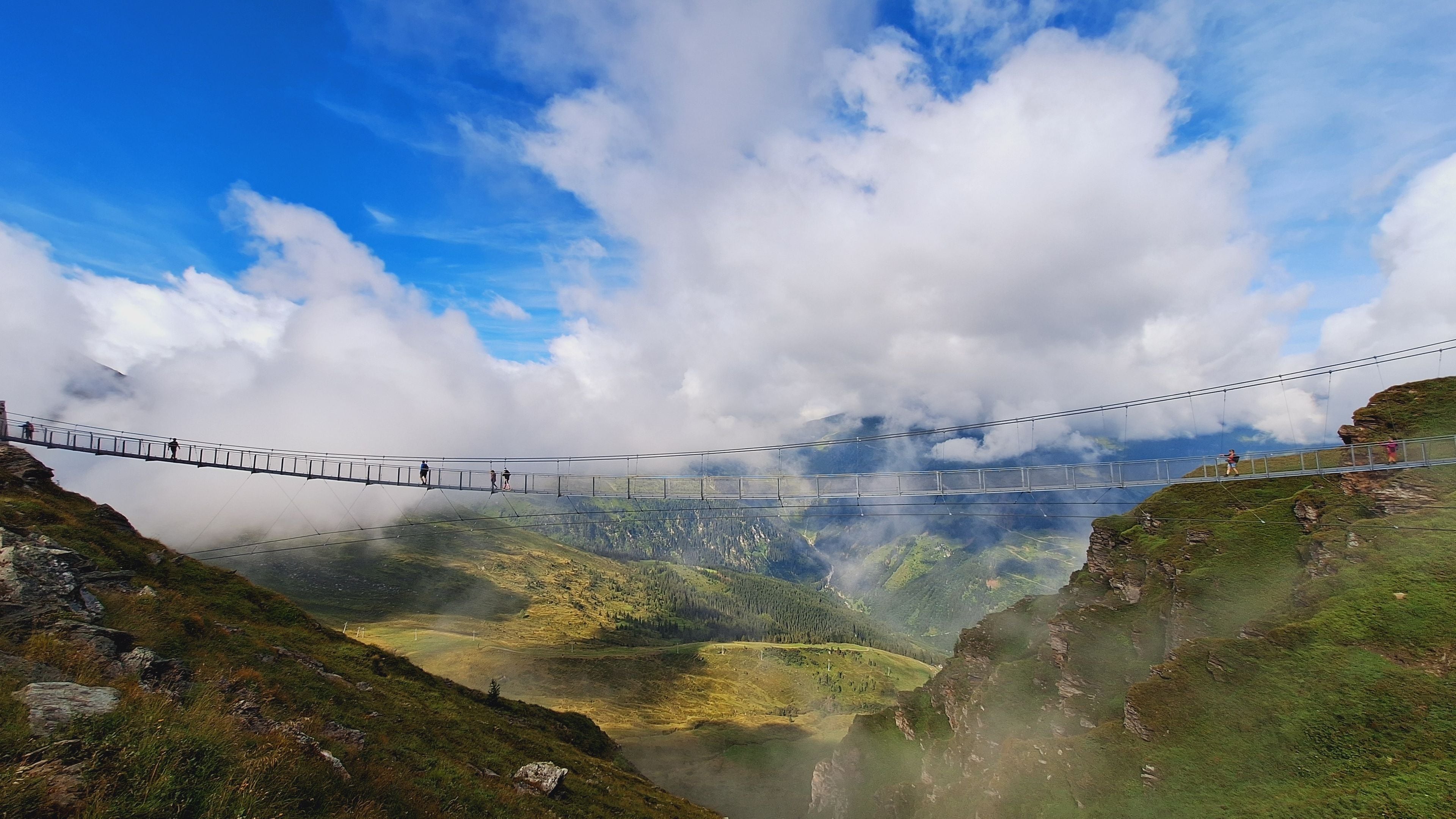 A suspension bridge over a valley with mountains in the background and clouds above.