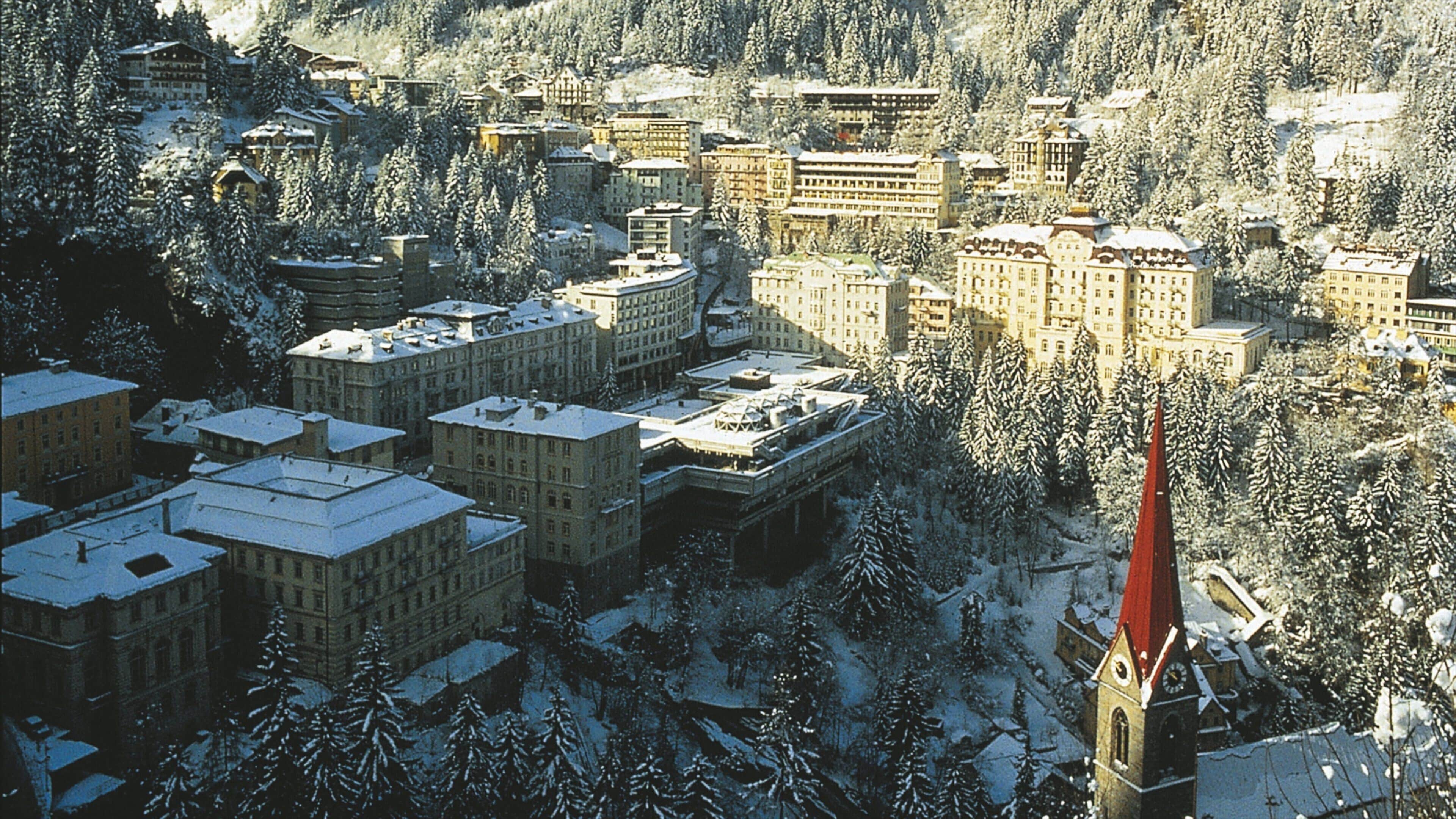 Uma cidade coberta de neve com vários edifícios, uma torre de igreja imponente e colinas arborizadas ao fundo.