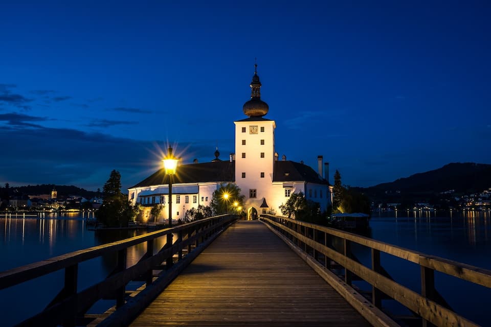 A wooden bridge leading to a white building with a tower and dome, surrounded by water and mountains.
