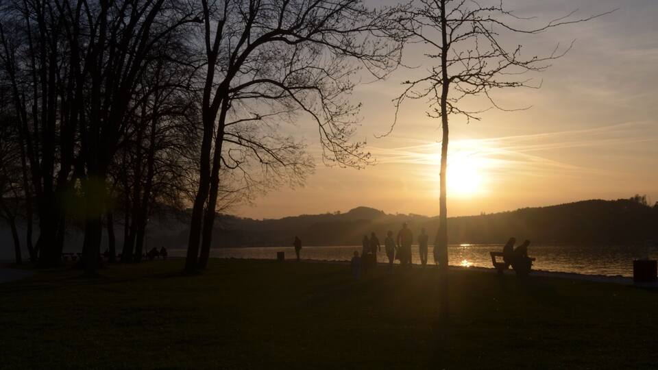 A sunset over a lake with trees and people on the shore.