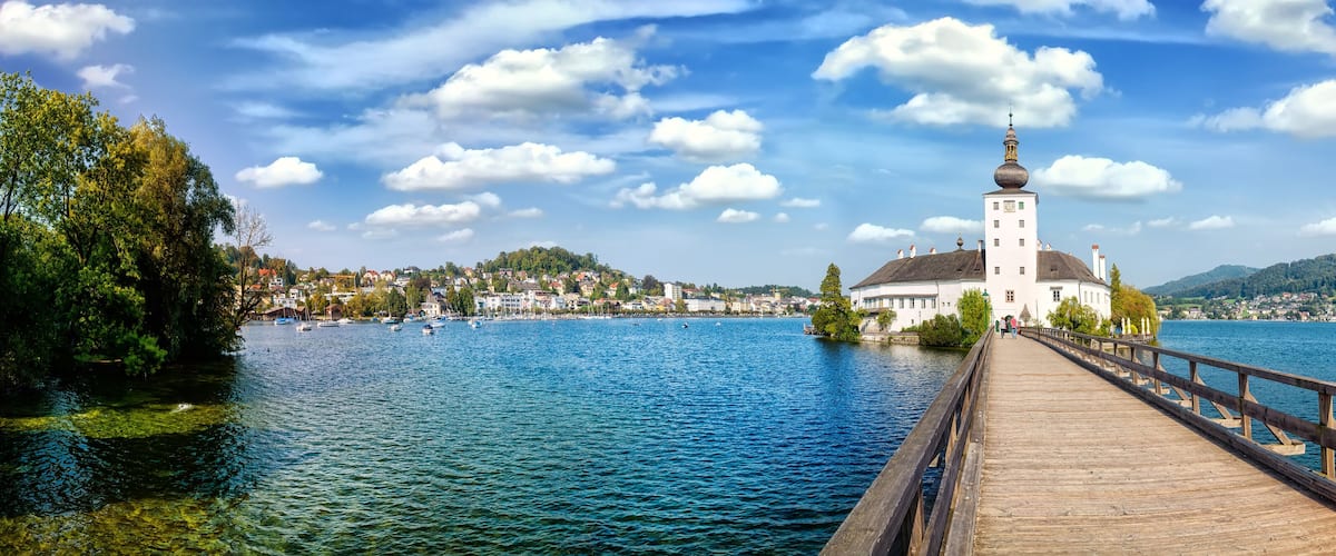 A wooden pier leading to a white church with a dome and bell tower, surrounded by a lake and a town.