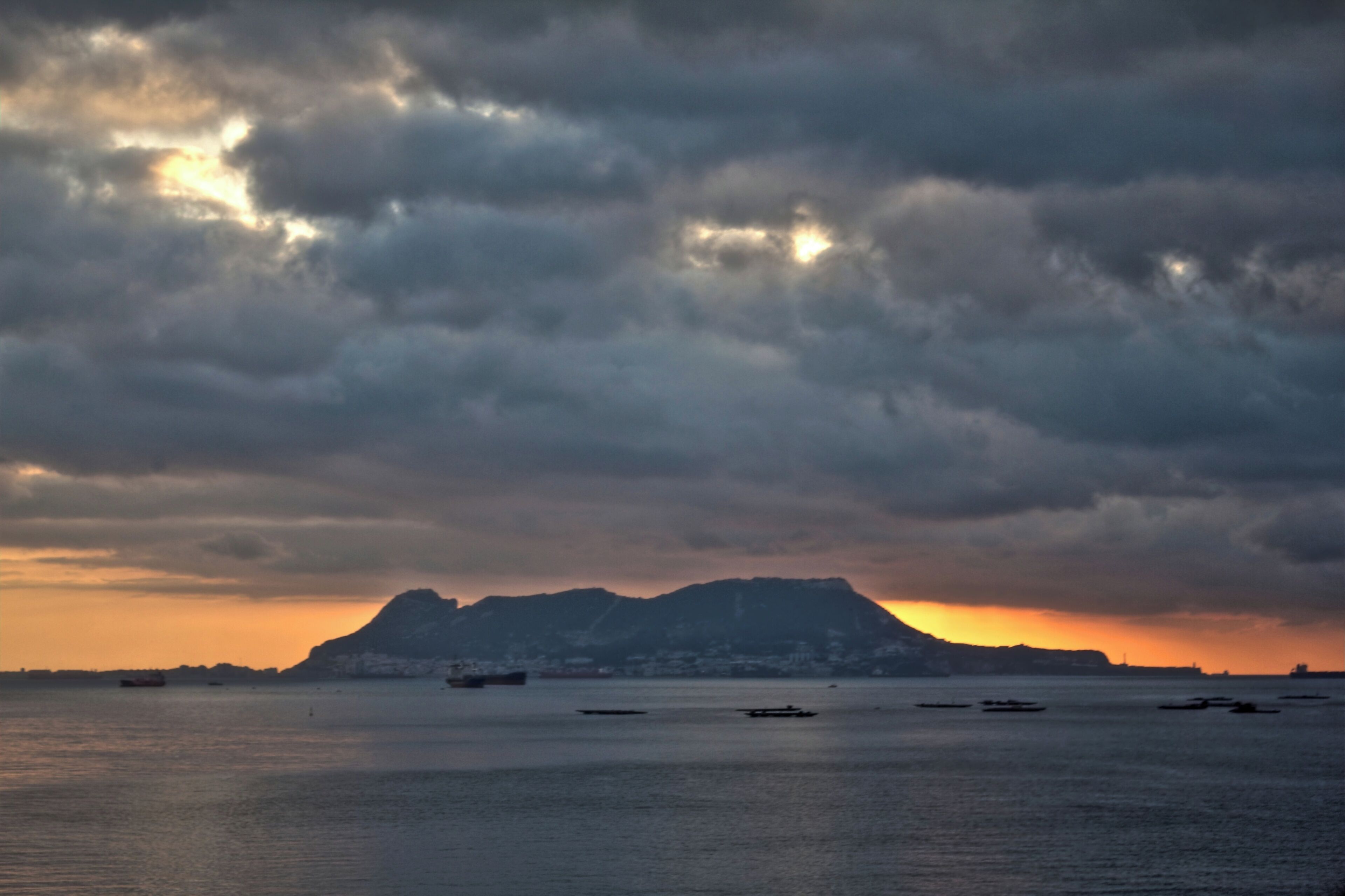 A sunset over a body of water with several boats, a distant mountain, and a cloudy sky.