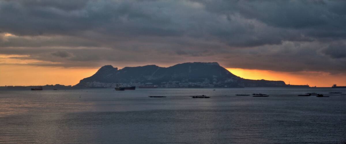 A sunset over a body of water with several boats, a distant mountain, and a cloudy sky.