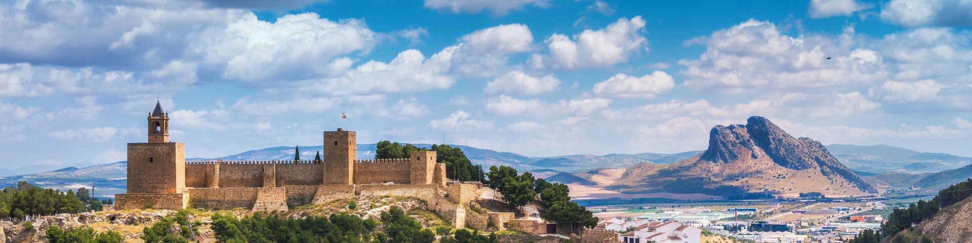 A historic fortress with a prominent tower, surrounded by lush greenery and a mountainous backdrop.