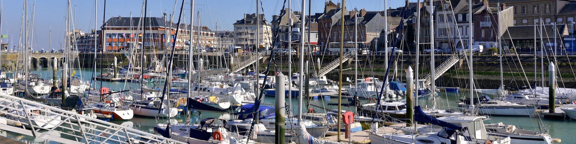 Un port de plaisance oĂč sont amarrĂ©s de nombreux voiliers, un pont en arriĂšre-plan et des bĂątiments le long de la rive.