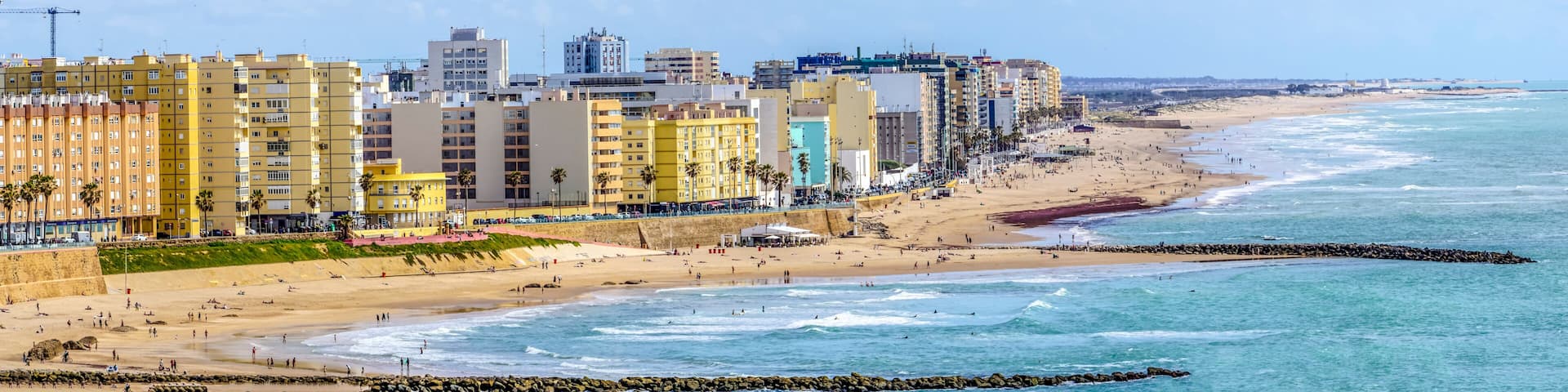 Wide panoramic view of the popular Playa de la Victoria in Cádiz, with its calm waters and golden sand in the sunlight, while the city buildings rise on the horizon