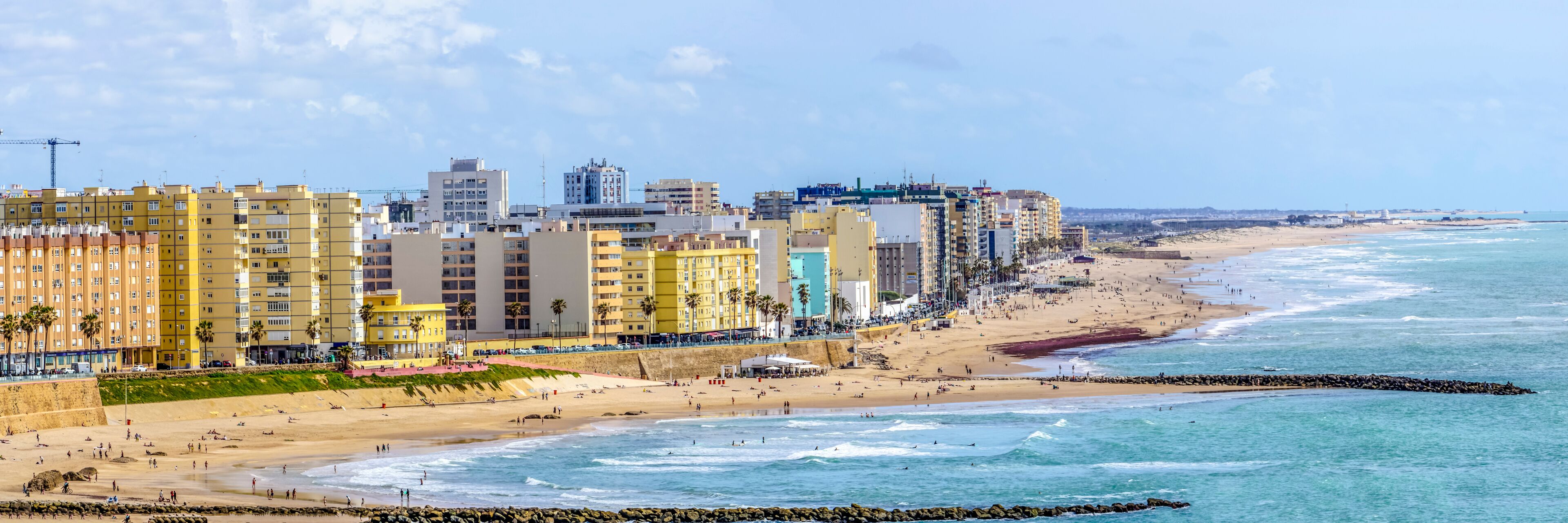 A coastal cityscape with a sandy beach, a rocky breakwater, and colorful high-rise buildings.