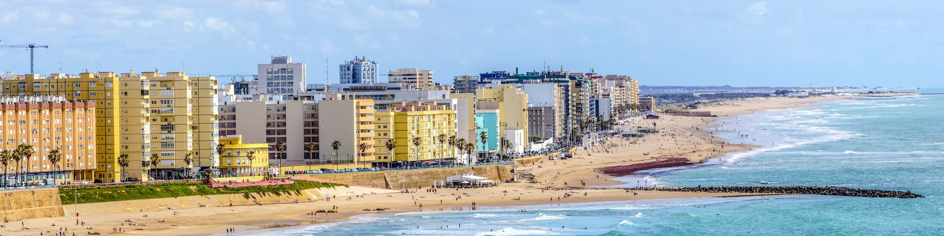 A coastal cityscape with a sandy beach, a rocky breakwater, and colorful high-rise buildings.