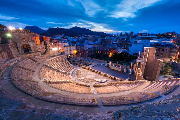 Un anfiteatro antiguo con escenario circular y asientos escalonados, rodeado de edificios históricos y montañas al fondo.