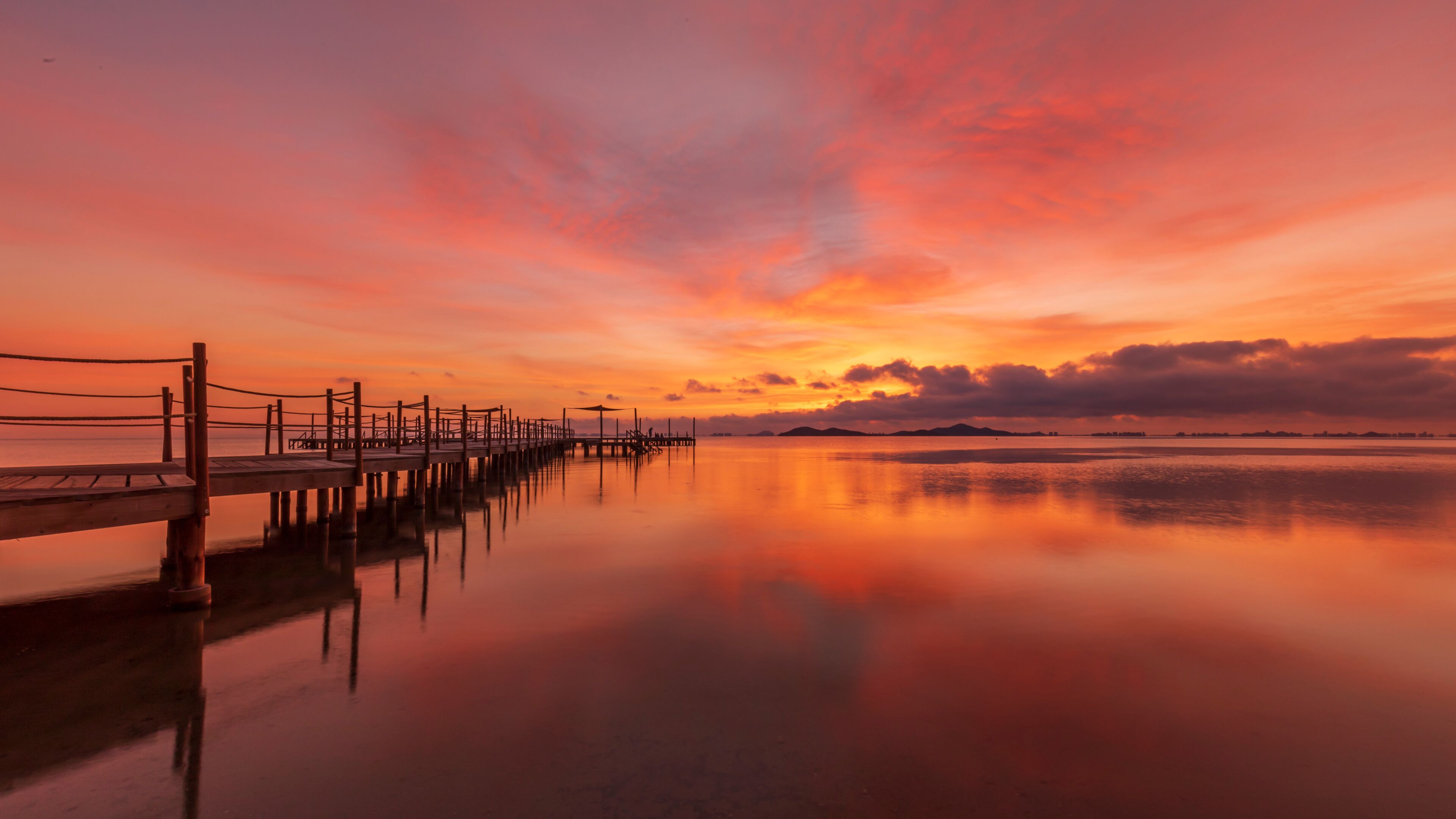 A wooden pier extending into calm waters, reflecting a sunset sky with orange and pink hues.