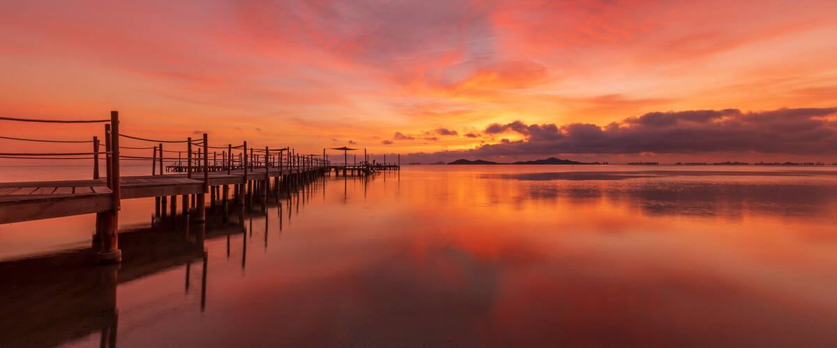 A wooden pier extending into calm waters, reflecting a sunset sky with orange and pink hues.