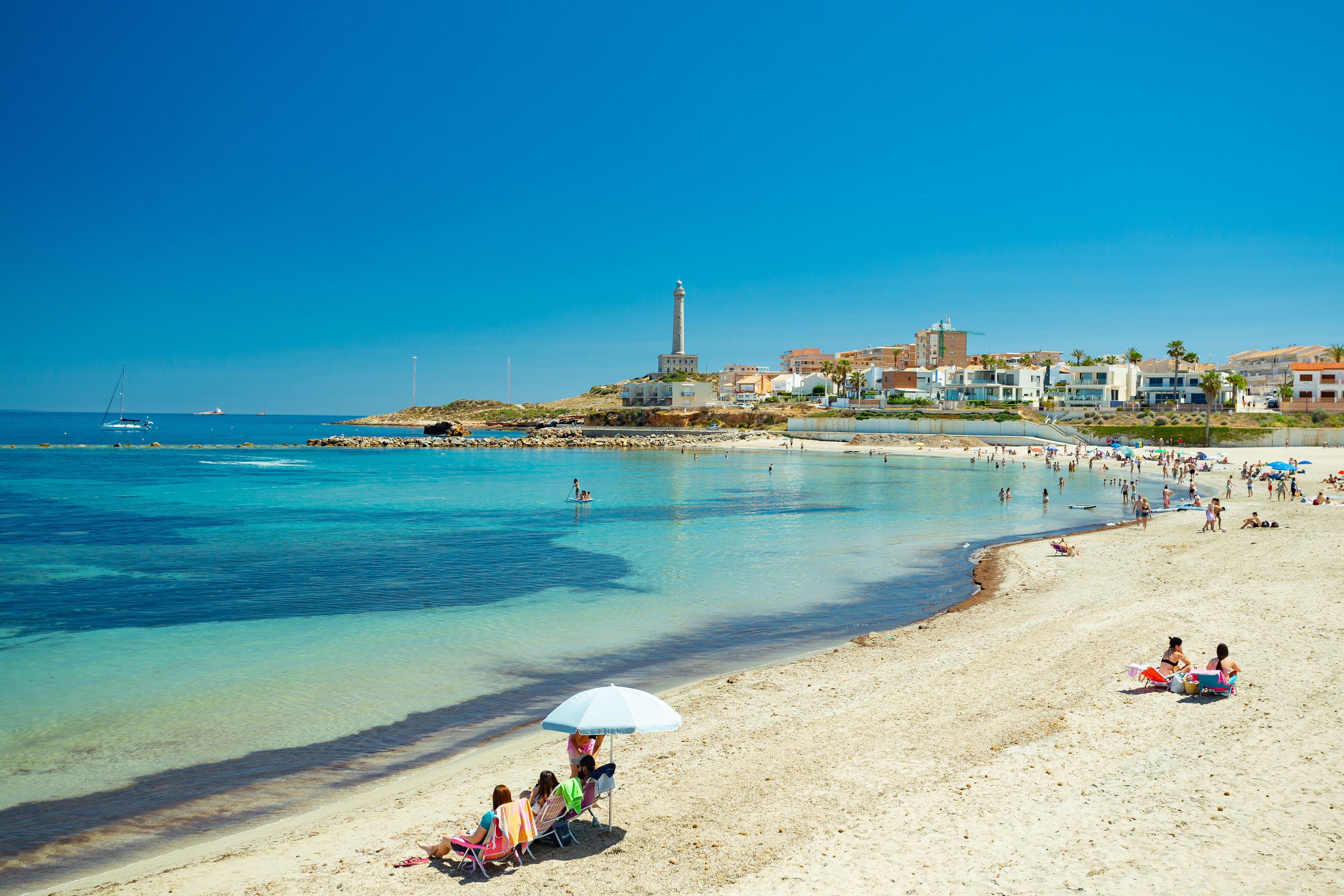 A sunny beach with clear blue water, a lighthouse, and buildings in the background.